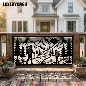 May include: A black metal decorative panel featuring a laser-cut silhouette of two hikers with backpacks and walking sticks on a winding trail. The scene includes pine trees, mountain peaks, and a sun. The text LUXLAYERED is visible at the top.