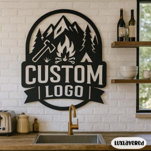 May include: Black metal wall art featuring a mountain scene with trees, a campfire, and an axe. The words "CUSTOM LOGO" are displayed prominently. The art is mounted on a white brick wall above a kitchen sink.