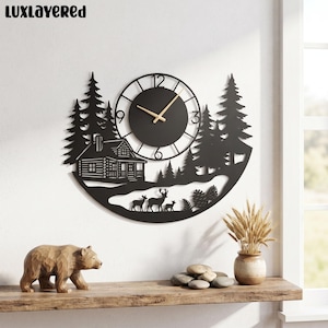 May include: A black metal wall clock featuring a laser-cut silhouette of a log cabin, pine trees, and a family of deer. The clock face has a circular center with gold-toned hands and numerals. The text LUXLAYERED is visible in the top left corner.