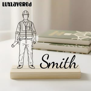 May include: A wooden desk nameplate with the name "Smith" in cursive. A black and white line drawing of a construction worker stands on the nameplate. The logo "LUXLAYERED" is in the top left corner.