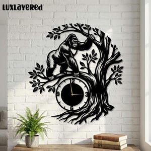 Puede incluir: Reloj de pared de metal negro con un gorila en una rama de árbol. La esfera del reloj está integrada en el diseño, con manecillas que indican la hora. El reloj está montado en una pared de ladrillos blancos. El texto "LUXLAYERED" está en la parte superior.