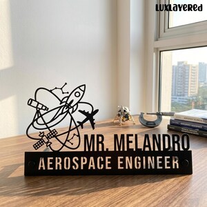 May include: A black metal desk sign featuring a cutout design of a rocket, satellite, airplane, and telescope. The text reads MR. MELANDRO and AEROSPACE ENGINEER in bold, capitalized letters. The sign is positioned on a wooden surface.