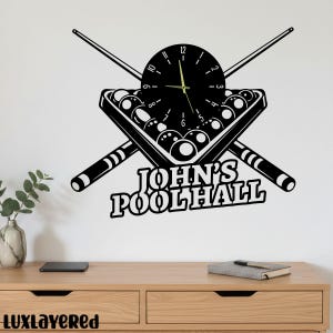 Könnte beinhalten: Schwarze Wanduhr mit Billard-Motiv. Die Uhr hat ein schwarzes Zifferblatt mit weißen Zahlen und Zeigern, einen Satz Billardkugeln, gekreuzte Billardqueues und den Text "JOHN'S POOLHALL".