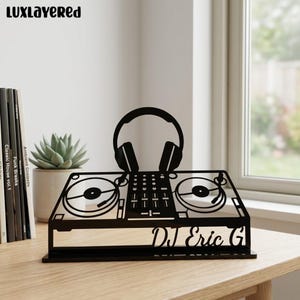 Puede incluir: Silueta negra de metal de una mesa de mezclas de DJ con auriculares. El diseño incluye dos tocadiscos, una mesa de mezclas y el texto "DJ Eric G". El producto se muestra sobre una superficie de madera.