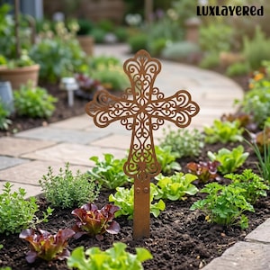 May include: A decorative, rusty-brown metal cross with an ornate, openwork design. The cross is staked into the soil of a garden bed filled with green and red leafy plants. The brand name "LUXLAYERED" is in the top right corner.