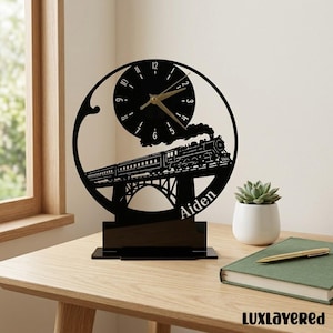 May include: Black laser-cut train clock with a circular design. The clock features a train on a bridge, with the name "Aiden" below. The clock hands are gold. The clock is on a black base and sits on a wooden table.