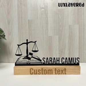 May include: A wooden desk sign featuring a black silhouette of scales of justice, a gavel, and an open book. The sign includes the name "SARAH CAMUS" and the words "Custom text" engraved on the wooden base. The logo "LUXLAYERED" is in the top right corner.