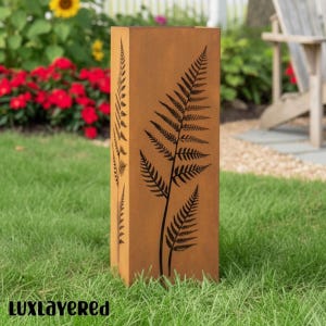Può includere: Un alto pilastro da giardino rettangolare in metallo color ruggine, caratterizzato da un design a foglia di felce tagliato al laser sui pannelli frontali e laterali. La superficie presenta una finitura ossidata e usurata.