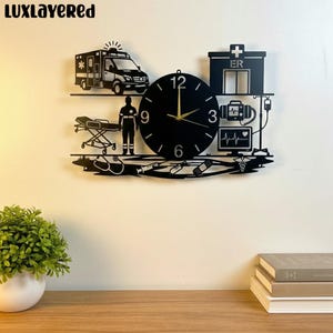 May include: Black metal wall clock with a medical theme. The clock features an ambulance, a stretcher, a medical professional, an ER building, and medical equipment. The clock hands are gold. The clock is mounted on a white wall.