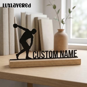 May include: A black metal silhouette of a discus thrower athlete positioned next to the text CUSTOM NAME, mounted on a rectangular light brown wooden base. The design features a clean, flat profile with a matte finish on the metal figure.