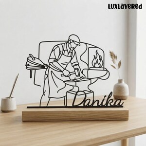 May include: A black metal wire art sculpture depicting a blacksmith working at an anvil with a bellows and fire in the background. The piece features the name Danika in cursive script and is mounted on a rectangular light wood base.
