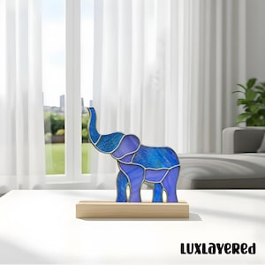 May include: A stained glass elephant sculpture in shades of blue and purple, mounted on a wooden base. The elephant has a detailed design with visible seams. The sculpture is displayed on a white table in front of a window.