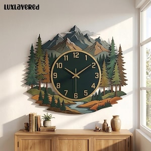 May include: A wall clock featuring a layered mountain and forest landscape with a dark green circular face, gold-colored numerals, and thin gold hands. The design includes pine trees, a river, and mountain peaks. The text LUXLAYERED is visible.
