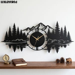 May include: Black metal wall clock with a mountain and forest silhouette design. The clock face is black with gold hands and numbers. The clock is surrounded by a detailed landscape scene, including trees and a lake.