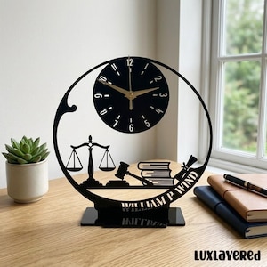 May include: Black laser-cut desk clock with a circular clock face and gold hands. The clock features a gavel, scales of justice, books, and a scroll design. The name "WILLIAM P WIND" is displayed on the base. The clock is on a wooden surface.