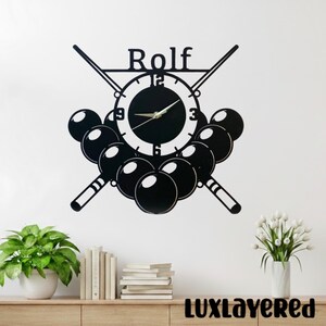 Könnte beinhalten: Schwarze Wanduhr mit Billard-Motiv. Die Uhr hat ein rundes Zifferblatt mit dem Namen "Rolf" darüber, gekreuzten Billardqueues und einer Ansammlung von Billardkugeln. Die Uhr ist an einer weißen Wand befestigt.