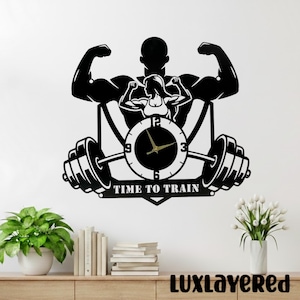 May include: Black metal wall clock with a fitness theme. The clock features a weightlifter silhouette, a woman silhouette, a barbell, and the words "TIME TO TRAIN". The clock hands are gold.