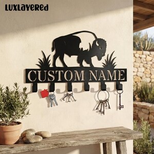 May include: A black metal wall-mounted key holder featuring a silhouette of a buffalo standing above grass. The horizontal bar displays the text CUSTOM NAME in capital letters and includes six hooks, with various metal keys hanging from them.