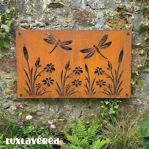 May include: A rectangular, rusty-orange metal wall art piece featuring two dragonfly silhouettes above a floral and cattail design. The artwork is mounted on a stone wall, with the text "luxlayered" visible at the bottom.
