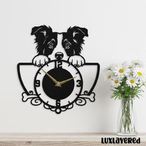 May include: Black metal wall clock featuring a dog design. The clock face is black with gold hands and numbers. The dog's head and paws are above the clock face. Decorative and functional wall art.