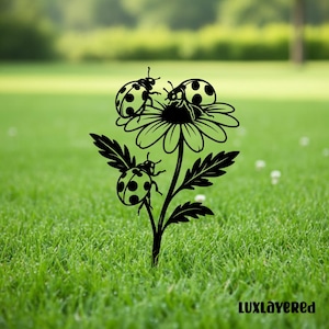 Può includere: Paletto da giardino in metallo nero raffigurante una margherita con tre coccinelle. Le coccinelle hanno macchie nere sul dorso. Il paletto è progettato per giardini o prati. Il testo "LUXlayered" è in basso a destra.