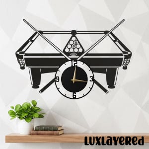Könnte beinhalten: Schwarz-weiße Wanduhr mit Billardtisch-Design und gekreuzten Queues. Das schwarze Zifferblatt hat goldene Zeiger und die Zahlen 3, 6, 9 und 12 sind sichtbar. Ein dekorativer Artikel für ein Spielzimmer.