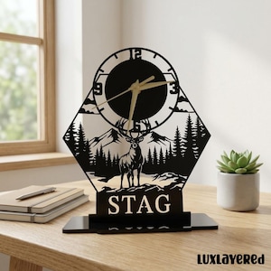 May include: Black laser-cut decorative clock featuring a stag design. The clock face is round with gold hands, set against a mountain and forest scene. The word "STAG" is displayed at the base. The clock is on a black stand.