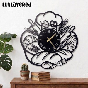 May include: Black metal wall clock with a baking theme. The clock face has Roman numerals and gold hands. The design includes a chef's hat, rolling pins, bread, a croissant, and a cupcake. The text "LUXLAYERED" is at the top.