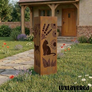 Puede incluir: Una escultura de jardín rectangular de metal envejecido con diseños recortados de gatos, flores y follaje. La obra de arte es de color marrón óxido cálido y se alza en posición vertical en un jardín cubierto de hierba. El texto "luxlayered" es visible en la esquina inferior derecha.