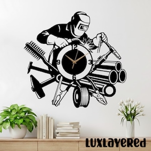 May include: Black and white wall clock featuring a welder with tools and pipes. The clock face is round with gold hands. The clock is surrounded by a hammer, brush, pliers, and other welding tools. The text "LUXLAYERED" is at the bottom.