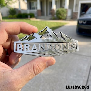 Puede incluir: Un emblema de metal plateado con un diseño de cordillera sobre el nombre "BRANDON'S" en un marco rectangular. El emblema se sostiene frente a una casa y un coche. La palabra "LUXLAYERED" está en la parte inferior.