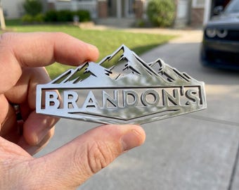 May include: A silver metal emblem featuring a mountain range design above the name "BRANDON'S" in a rectangular frame. The emblem is held up in front of a house and car. The word "LUXLAYERED" is at the bottom.