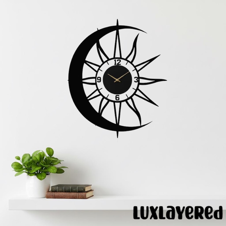 May include: Black metal wall clock featuring a sun and crescent moon design. The clock face has gold-colored hands and numerals. The clock is mounted on a white wall. The text "LUXLAYERED" is at the bottom.