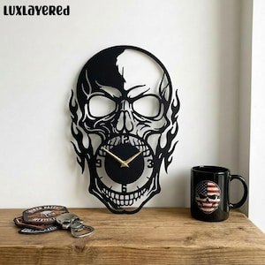 Könnte beinhalten: Schwarze, schädelformige Wanduhr mit goldenen Zeigern, Flammen und einem kreisförmigen Zifferblatt. Eine schwarze Tasse mit einem Totenkopf und amerikanischem Flaggen-Design, ein Totenkopf-Flaschenöffner und Aufnäher befinden sich auf einer Holzoberfläche. Der Markenname "LUXLAYERED" steht oben.