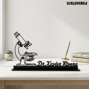 May include: A black laser-cut desk sign featuring a silhouette of a microscope and the name Dr. Xavier Rivera in a cursive script font. The sign is mounted on a rectangular base and is positioned on a white surface with a pen and books nearby.