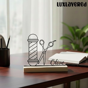 Puede incluir: Un accesorio de escritorio con temática de barbería en blanco y negro. El diseño presenta un poste de barbero y unas tijeras, montados sobre una base de madera. El nombre "Olive" está escrito en cursiva. El fondo incluye un escritorio, un portalápices y un cuaderno.