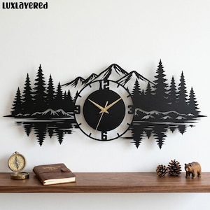 May include: A black wall clock featuring a laser-cut silhouette of a mountain range and pine forest reflected in water. The circular clock face has gold-colored hands and numerals 3, 6, 9, and 12. The text LUXLAYERED appears in the top left corner.