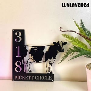 May include: Black and white metal cow silhouette with the address numbers 318 and the text "PICKETT CIRCLE". The sign has a decorative fern and the text "LUXLAYERED" in the upper right corner.