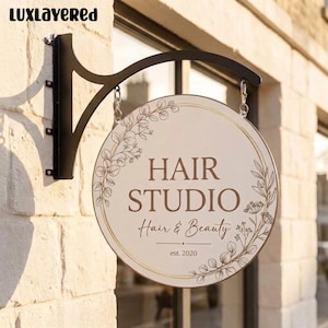 May include: A round, off-white hanging sign with a black metal bracket. The sign features brown text reading HAIR STUDIO Hair & Beauty est. 2020, surrounded by a delicate gold-colored circular frame and botanical line art illustrations of leaves.