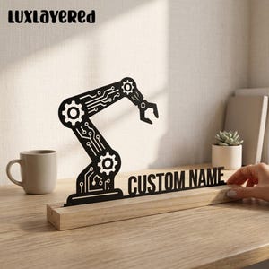 Könnte beinhalten: Eine schwarze, lasergeschnittene Silhouette eines Roboterarms mit integrierten Leiterplattenmustern und Zahnradmotiven, montiert auf einem rechteckigen, hellen Holzsockel. Der Sockel enthält den Text CUSTOM NAME in fetten schwarzen Buchstaben.