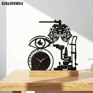 May include: A black metal desk clock featuring an optometry theme with a phoropter, a slit lamp, and a stylized eye. The clock face is circular, includes the name CHRISTINA, and has gold-colored hands, all mounted on a rectangular wooden base.