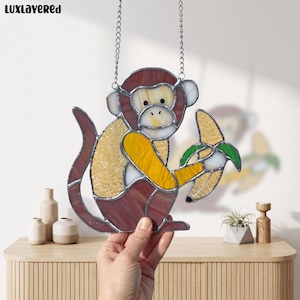 Peut inclure: Un attrape-soleil en vitrail en forme de singe tenant une banane, composé de pièces de verre marron, jaune et blanc assemblées par des lignes de soudure en métal argenté. L'objet inclut une chaîne argentée et le texte LUXLAYERED dans le coin.