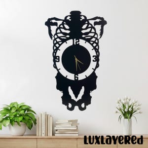 May include: Black metal wall clock with a unique design. The clock features a circular face with gold hands and a decorative frame. The clock is mounted on a white wall above a wooden shelf with plants and books. The text "LUXLAYERED" is at the bottom.
