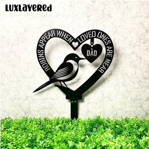 Könnte beinhalten: Schwarzer Metall-Gartenstecker mit einem Vogel, der auf einem Ast in einem herzförmigen Rahmen sitzt. Der Rahmen trägt den Text "ROBINS APPEAR WHEN LOVED ONES ARE NEAR" und ein herzförmiges Etikett mit der Aufschrift "DAD". Der Markenname "Luxlayered" befindet sich oben links.