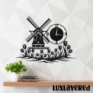 Könnte beinhalten: Schwarze Metall-Wanduhr mit Windmühlendesign und Ziffernblatt. Die Uhr hat einen schwarzen Rahmen und goldene Zeiger. Die Windmühle ist von Tulpen umgeben. Die Uhr ist an einer weißen Wand über einem Holzregal befestigt.
