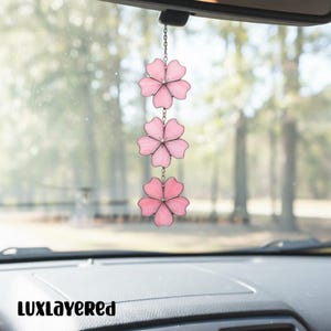May include: A car charm featuring three pink flower pendants. Each flower has five petals and is connected by a silver chain. The charm hangs from a car's rearview mirror. The text "LUXLAYERED" is at the bottom.