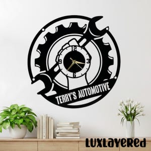 May include: Black metal wall clock with a gear and wrench design. The clock face is round with gold hands and numbers. The text "TERRY'S AUTOMOTIVE" is displayed on the clock. The clock is mounted on a white wall.