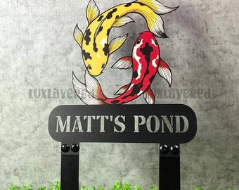 Może przedstawiać: Czarny metalowy palik ogrodowy z napisem "MATT'S POND" i dwoma karpiami koi. Jeden karp jest żółty z czarnymi plamami, a drugi czerwony z czarnymi oznaczeniami. Karpie znajdują się nad znakiem.