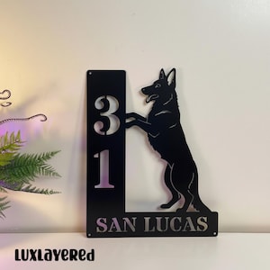 Puede incluir: Letrero de dirección de metal negro con los números "31" y el nombre "SAN LUCAS". Una silueta de un perro pastor alemán está de pie junto a los números. El letrero está diseñado para uso en exteriores.