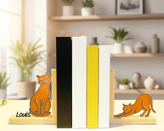 Puede incluir: Sujetalibros con forma de gato naranja, con base de madera, que sostienen libros negros, blancos y amarillos. Un gato está sentado, el otro se estira. El nombre "Louis" está escrito en la base.